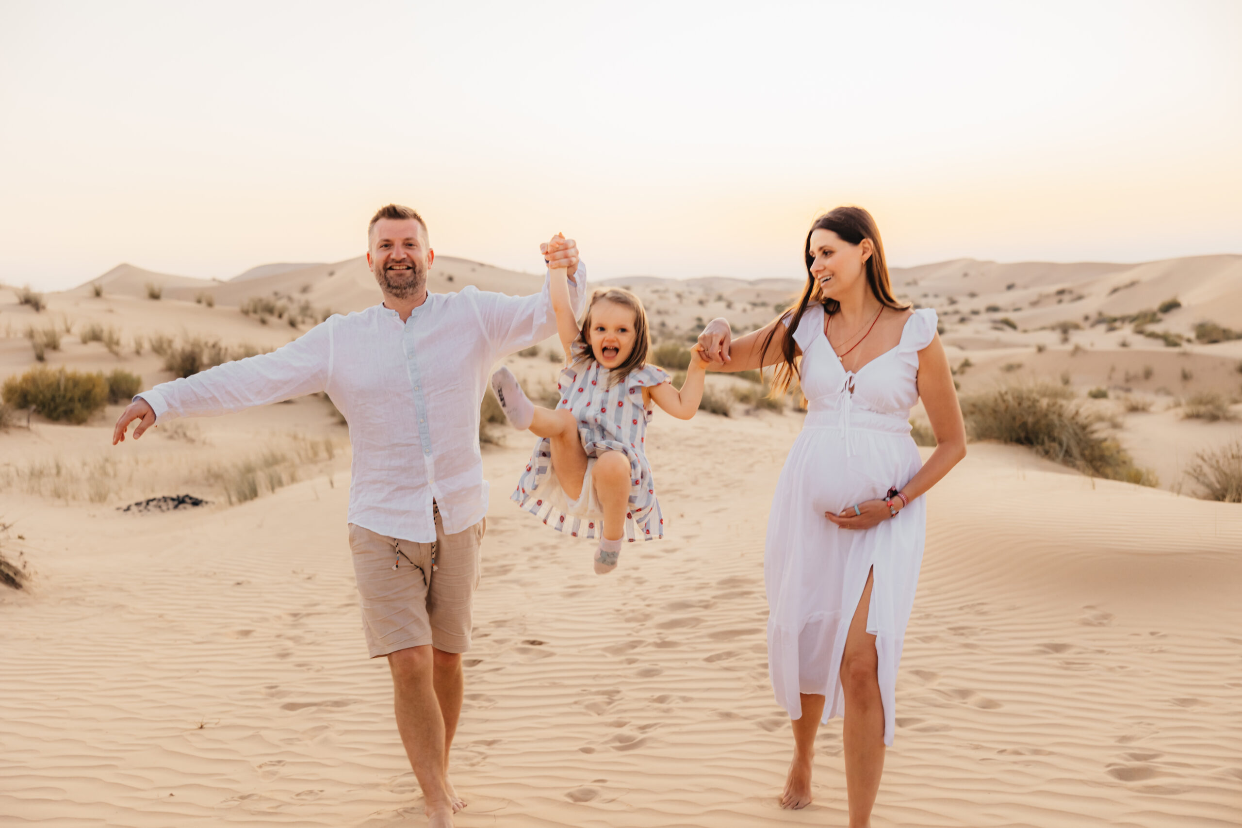 Parents lifting their daughter into a jump while the mother holds her baby bump during a joyful desert maternity family session in Abu Dhabi.