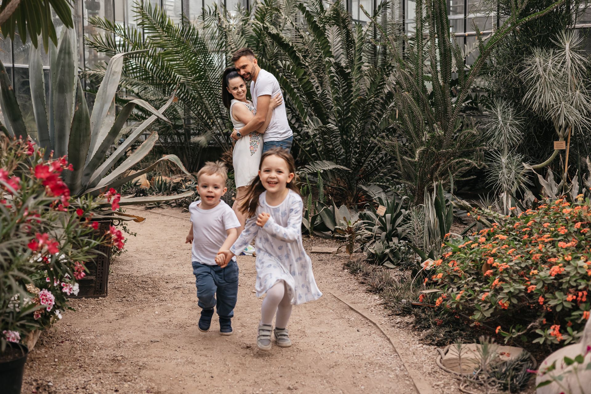 Parents embracing in the background while watching their children run toward the camera during a joyful family session in Umm Al Emarat Park in Abu Dhabi.